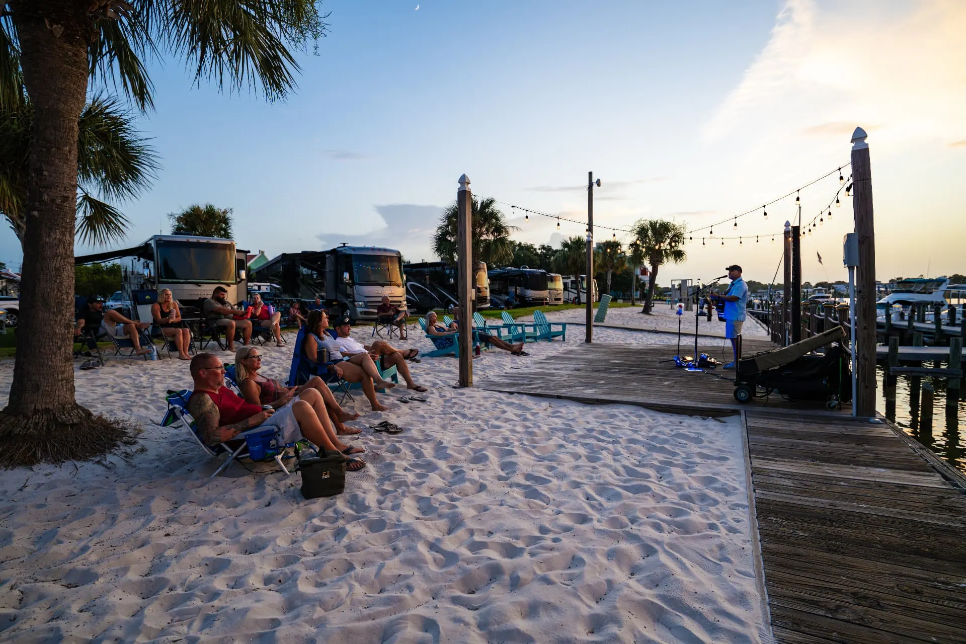 Guests watching live music on the beach at sunset beside the marina